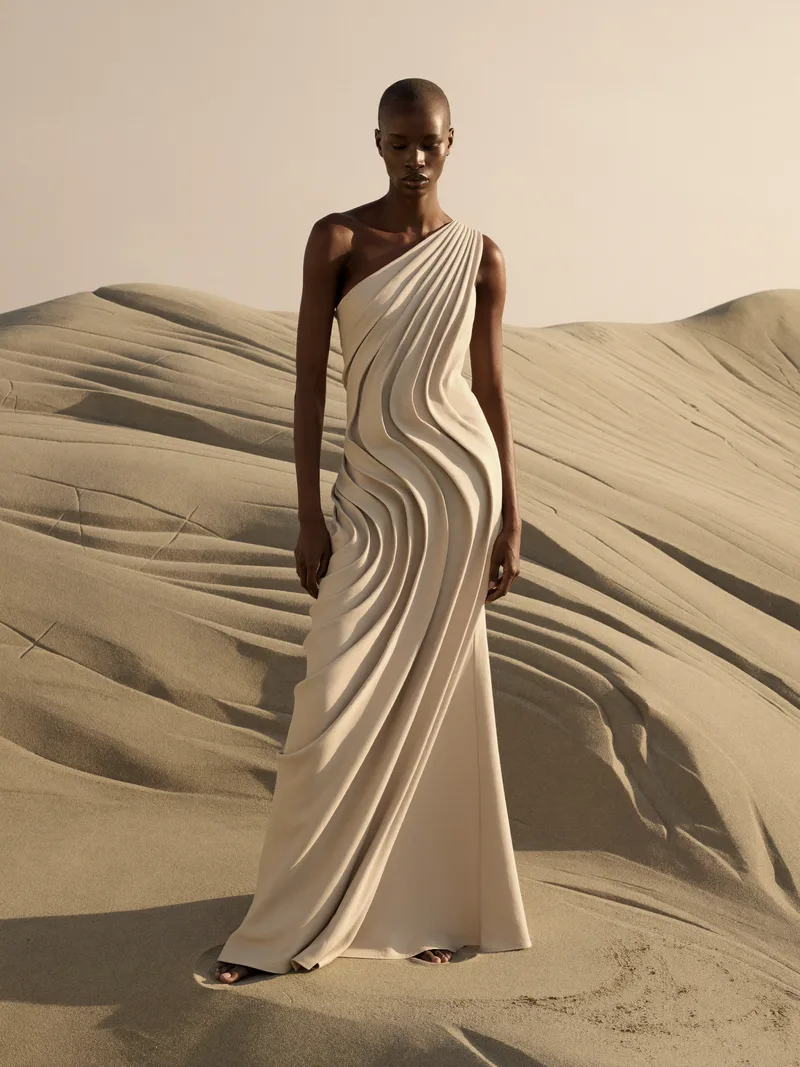 Look I — a model in a floor-length ecru gown of spiral pleats, standing in a sculpted sand dunescape